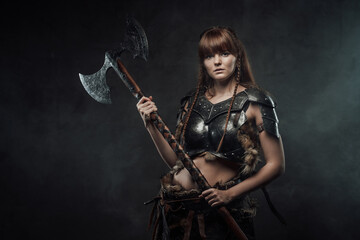 Barbaric beutiful woman from north armed with two handed axe and with brown hairs poses in dark foggy background.