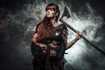 Barbaric female viking in light armour with brown hairs poses in dark smokey background holding two...