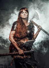 Armed with two huge axes scandinavian female warrior with brown hairs in dark armour poses in smokey background.
