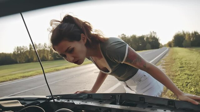 Sexy asian woman wearing white shorts and T-shirt looking under the hood of a broken car. Young pretty brunette trying to repair her automobile on the side of the road. Concept of promblem