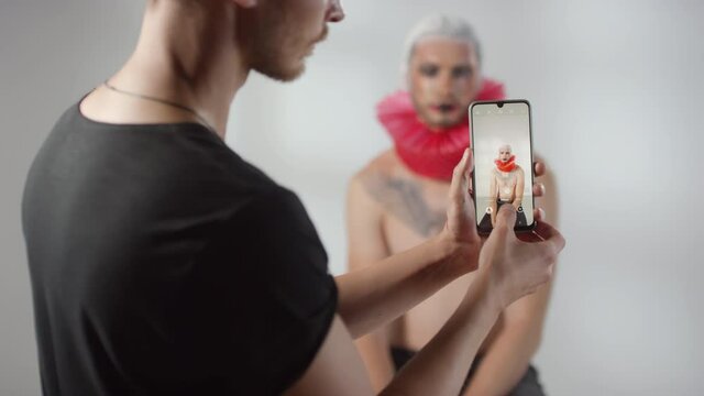 Medium shot of man holding mobile phone and taking photo of shirtless male model with dramatic make-up and red ruff around his neck