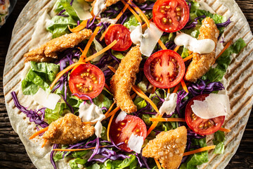 Top view of open tortilla with chicken nuggets, chery tomatoes, red cabbage, salad, carrot stripes and parmesan shavings