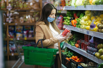 Happy woman in face mask taking fresh cherry tomatoes while standing at groceries in supermarket. Beautiful young girl with food basket choosing red tomatoes by stand with vegetables. Quarantine