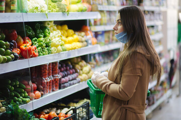 Happy woman in protective mask taking fresh vegetables while standing by groceries in supermarket. Beautiful young girl with food basket choosing food by stand with vegetables. Christmas purchases