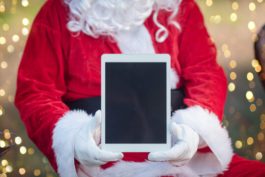 Santa Claus using tablet pc computer for communication. Christmas tree and beautiful decor bokeh background. Happy busy holiday and shopping time.