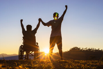 Silhouette of joyful disabled man in wheelchair raised hands with friend at sunset