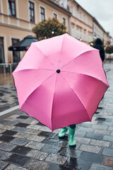 Child hiding behind big pink umbrella walking in a downtown on rainy gloomy autumn day