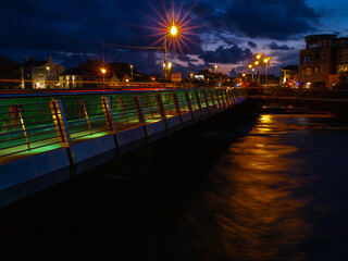 Fototapeta na wymiar Night shot of Wolfe Tone Bridge in Galway city, Ireland, Blue sky, warm orange lights reflects in river Corrib. Pronounced lens star-burst.