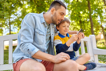 family, fatherhood and technology concept - happy father and little son with smartphone sitting on bench at summer park
