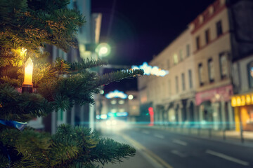 Night scene, candle on a fur tree in focus, town decorated and illuminated for Christmas out of focus. Festive season concept