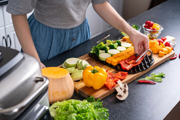 Woman lays sliced ​​vegetables on grill grate, in the home kitchen. Blured foreground.