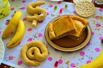 Wooden brunch table with a white tablecloth with pink polka dots, empanada, bananas, rice cakes, at sign or dollar symbol breads, gummy bears and oat flakes