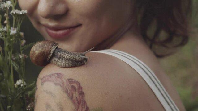 Woman holding bouquet of wild flowers or daisies in her hands. Close-up of snail slowly crawling along shoulder of young girl. Snail leaves muscus on skin. Concept of nature, cosmetology, spa, beauty.