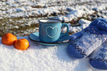 Obraz na płótnie Canvas Blue mug with a heart pattern, mittens and two orange tangerines on the snow, side view-the concept of a pleasant winter holiday