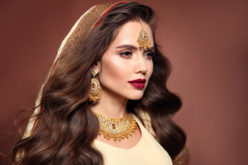 Beautiful woman with long wavy hair style and bright makeup. Young hindu brunette model with kundan golden jewelry set isolated on brown studio background. - 396977116