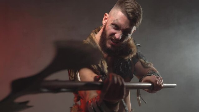 Angry and violent viking warrior armed with axe and dressed in armour with fur moves forward and attacks and then moves back in dark and smokey background.