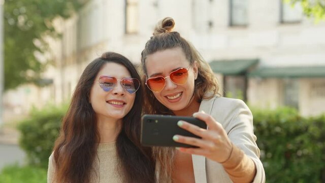 Two young smiling hipster blond women taking selfie self portrait photos on smartphone. Pretty stylish girls on street background. Positive emotions and love. LGBTQI, Pride Event, LGBT Pride Month