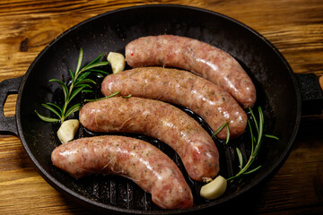 Raw sausages ready for preparation with rosemary, garlic and spices in cast iron grill frying pan on wooden table