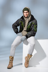 full length of trendy man in hat, gloves and parka sitting with hand on hip on white cube on grey © LIGHTFIELD STUDIOS