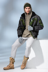 full length of trendy man in hat, gloves and parka sitting on white cube on grey © LIGHTFIELD STUDIOS