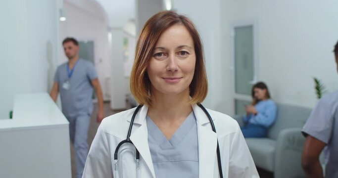 Close up portrait of happy Caucasian beautiful female physician with stethoscope in white coat standing in clinic, looking and smiling to camera. Woman doctor at workplace. Healthcare concept