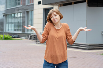 An adult redhead woman stands in front of a closed building of a business center spreading her arms and shrugging her shoulders in a questioning gesture. She doesn't know what will happen next