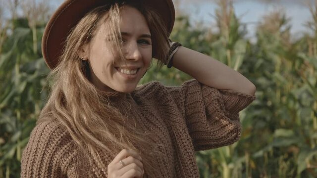 Close up side or profile portrait of blonde hipster smiling and laughing sincerely and broadly while looking into camera. Girl with blonde hair in hat standing in middle of corn field or meadow.