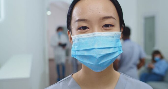 Close Up Portrait Of Asian Happy Young Beautiful Female Hospital Practitioner Wearing Mask While Standing In Clinic Hall And Smiling At Camera. Coronavirus Pandemic. Healthcare Worker. Covid-19