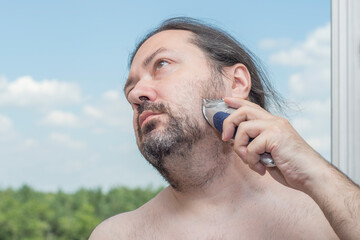 An ordinary middle-aged adult man with ponytail hair shaves his beard with an electric trimmer. Man on a light background.