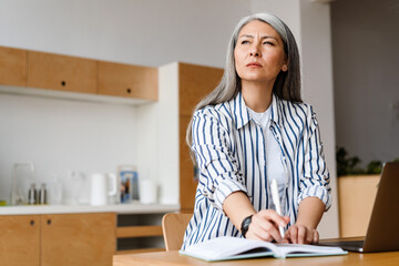Serious white-haired mature woman writing down notes working with laptop
