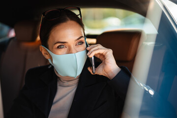 Pleased businesswoman wearing face mask talking on mobile phone in car