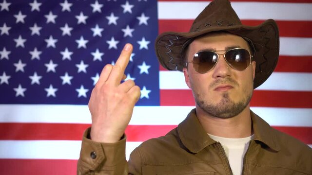 Portrait of a serious man in a cowboy hat, leather jacket and sunglasses on the background of the American flag, the man shows an indecent sign, a phallic symbol.