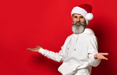 Fototapeta na wymiar Portrait of a handsome senior bearded man, in a white hoodie and santa hat, stands on a red background with his hands spread apart