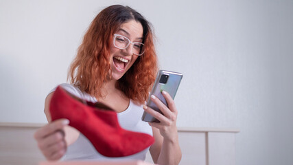 Fototapeta premium Online shopping using the application on the phone. Happy woman unpacks shoes and holds a smartphone. The expectation and reality of the purchase is consistent