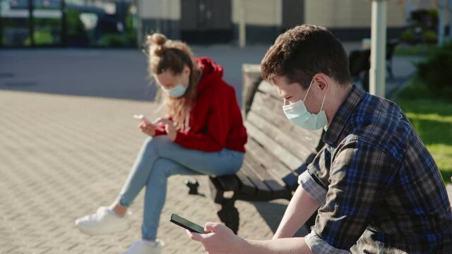 Social distancing. Young people sitting on different benches at safe distance in protective masks and talking. Daily routine after quarantine. Communicating with each other using modern technology.