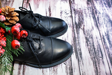 Black leather shoes and Christmas decor on wooden background. Pair of stylish women's boots and spruce twig with Christmas toys on the laminate floor. Leather shoe care, custom tailoring, shoe repair