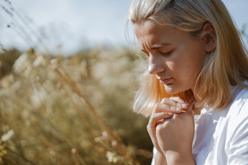 A teenage girl closed her eyes while praying in a field. Hands folded in prayer faith, spirituality and religion concept