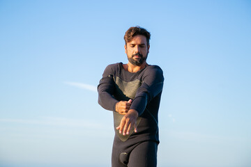 Focused sportsman putting on wetsuit for surfing on ocean beach. Low angle, blue sky background. Active lifestyle concept