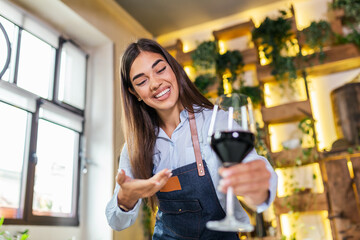 Young beautiful waitress wearing apron holding a glass of red wine in one hand serving a customer in a rustic restaurant. Sommelier recommended wine in restaurant.
