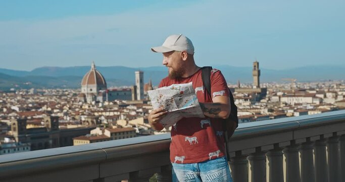 Attractive tourist man in white cap trying to identify location on the map. Handsome bearded guy exploring Italian city from viewpoint at sunset. Concept of Holidays, Vacations and Travel in Europe.