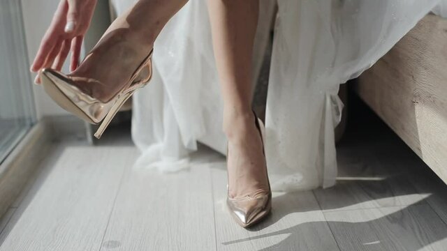 Close-up of legs of a young bride putting on beautiful gold high-heeled shoes while preparing for a wedding ceremony