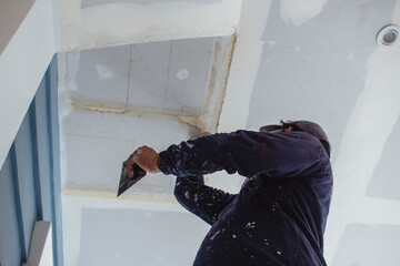A man applies putty to a ceiling of an almost complete home. Wearing clothes full of paint...