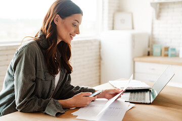 Joyful woman using cellphone while working with laptop and papers