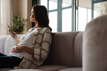 Happy charming pregnant woman using mobile phone while sitting on sofa