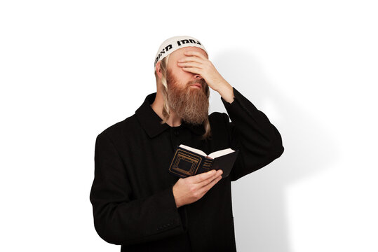Religious Jewish man holding Siddur in his hand covering eyes with hand isolated on white background. Bearded Jew hasid with sidelocks in white kippah reads prayer. Reading Kriyat Shema.