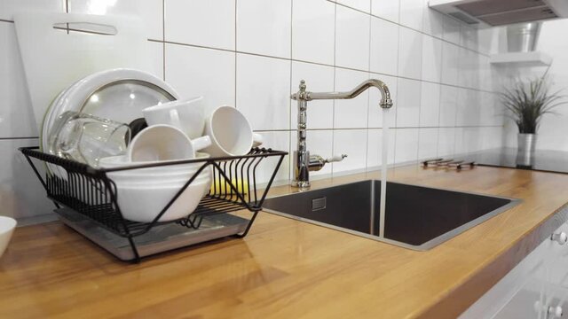 Water flowing out of stainless steel tap. Dish rack holds dishes against wooden countertop, white wall tiles, sink and faucet. Budget lightweight dish drainer with drain board at scandinavian kitchen