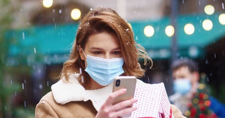 Beautiful Caucasian woman with blond hair stands on snowy street in medical mask holding gift and writing message on smartphone.