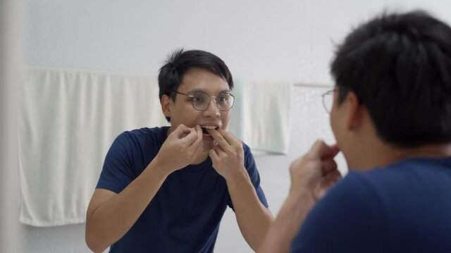 asian man using dental floss removing food particles that left on teeth off in mirror at white bathroom.