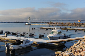 A pier on a harbour on Vättern lake in Gränna in Sweden on a sunny winter day with white snow, blue sky, clear air and water, a lighthouse and yachts