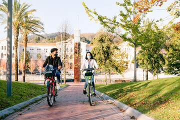 front of two young man and woman riding along a bicycle path with a sharing electric bicycle in a beautiful park with many trees at sunset wearing a face mask for the 2020 covid19 coronavirus pandemic